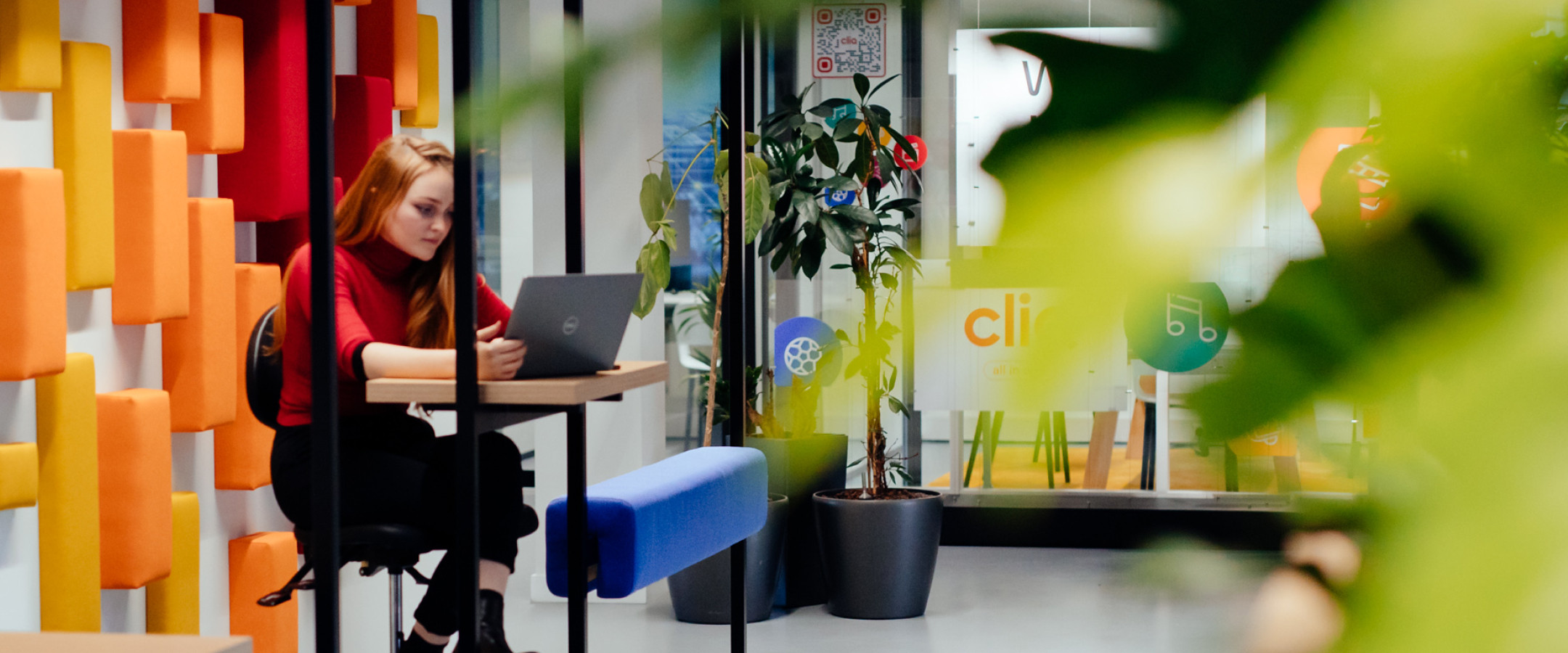 Woman working at her desk in front of brightly colored wall. A blurry plant is in the foreground.